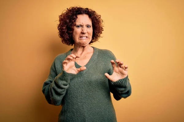 Middle age beautiful curly hair woman wearing casual sweater over isolated yellow background disgusted expression, displeased and fearful doing disgust face because aversion reaction. With hands raised