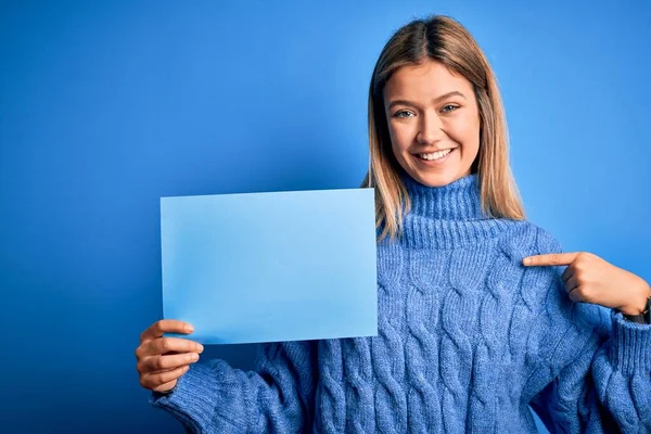 Young beautiful woman holding banner standing over isolated blue background with surprise face pointing finger to himself