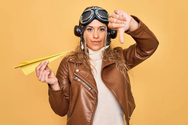 Beautiful blonde aviator woman wearing vintage pilot helmet whit glasses holding paper plane with angry face, negative sign showing dislike with thumbs down, rejection concept