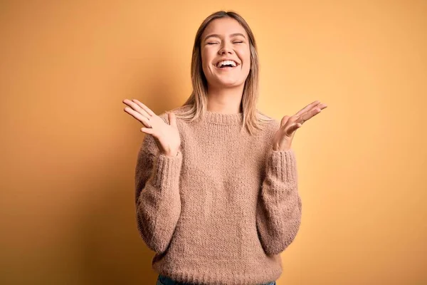 年轻美丽的金发女人穿着棕色毛衣 披着黄色的孤立的背景 疯狂而疯狂地庆祝成功 举手表决 紧闭双眼兴奋地尖叫着 赢的概念 — 图库照片