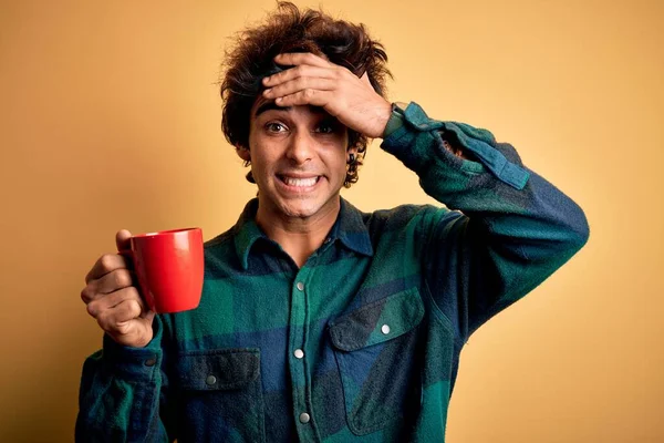 Young handsome man drinking cup of coffee standing over isolated yellow background stressed with hand on head, shocked with shame and surprise face, angry and frustrated. Fear and upset for mistake.