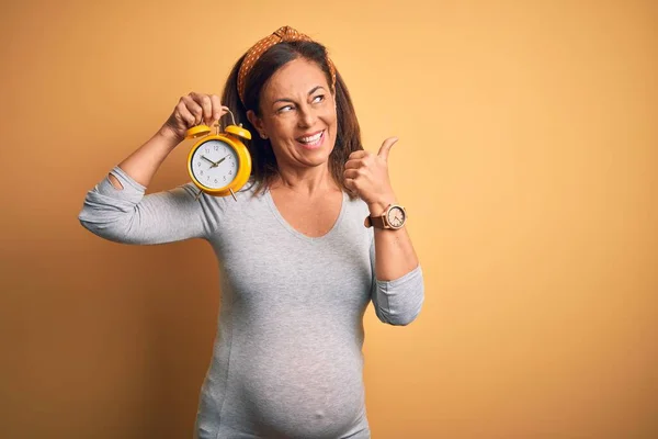中年妊娠中の赤ちゃんを期待します妊娠ホールディング目覚まし時計ポインティングとととともに親指で側まで幸せな顔笑顔 — ストック写真