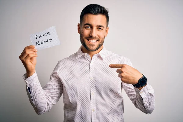 Young business man holding fake news paper over isolated background with surprise face pointing finger to himself