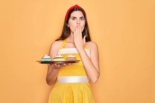 Young beautiful pin up woman wearing 50s fashion vintage dress holding tray with cupcakes cover mouth with hand shocked with shame for mistake, expression of fear, scared in silence, secret concept