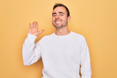 Mavi gözlü genç adam sarı arka planda dikilmiş mutlu ve güler yüzlü dostça bir jest yapıyor.