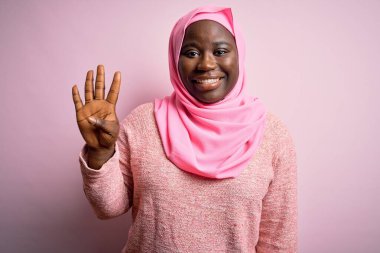 Genç Afro-Amerikan artı cüsseli kadın tecrit edilmiş pembe arka plan üzerine Müslüman başörtüsü takıyor ve parmaklarıyla dört numarayı gösterirken kendinden emin ve mutlu gülümsüyor..