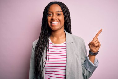Genç Afro-Amerikan iş kadını pembe izole edilmiş arka planda duruyor, yüzünde kocaman bir gülümsemeyle, kameraya el ile işaret ediyor..