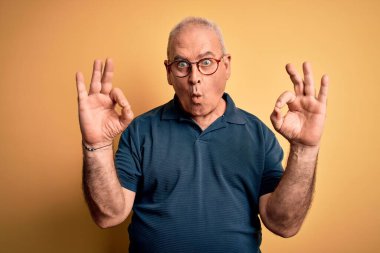 Sarı arka plan üzerine gündelik polo ve gözlük takan orta yaşlı yakışıklı adam şaşırmış ve şaşırmış görünüyor. Çılgın ifade