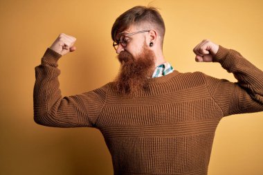 Sarı arka planda sakallı ve kışlık süveter giyen yakışıklı kızıl saçlı yakışıklı adam, gururla gülümseyen kol kaslarını gösteriyor. Fitness konsepti.