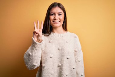 Mavi gözlü genç bir kadın sarı arka planda dikilen ve üç numaralı parmaklarıyla gülümseyerek mutlu ve kendinden emin bir şekilde dikiliyor..