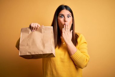 Elinde paket poşeti tutan genç bir kadın sarı arka plan üstü örtülmüş ağız hata yüzünden utançla şok olmuş, korkunun ifadesi, sessizlikten korkmak, gizli konsept