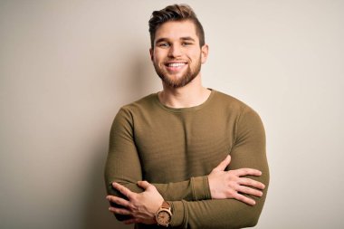 Sakallı, mavi gözlü, beyaz arka planda yeşil süveter giyen, kollarını kavuşturup kameraya bakan genç, sarışın bir adam. Pozitif kişi.