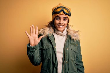 Genç Afrikalı Afro-Amerikan kayakçı kız kar sporu giysisi ve kayak gözlüğü takıyor ve parmaklarını 5 numarayı göstererek gülümsüyor ve mutlu..