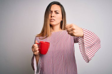 Mavi gözlü güzel sarışın kadın beyaz arka planda kırmızı kahve fincanı içerken sinirlenmiş ve sinirlenmiş öfkeyle bağırıyor, çıldırmış ve el kaldırarak bağırıyor.