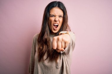 Genç, güzel, esmer bir kadın pembe arka planda dikilmiş sana kızgın ve kızgın bir şekilde kameraya kızgın olduğunu gösteriyor.