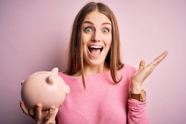 Genç, güzel, kızıl saçlı, kumbarayı soyulmuş pembe arka planda tutan mutlu ve heyecanlı, kazanan yüz ifadesi zaferi büyük bir gülümsemeyle kutluyor ve ellerini kaldırıyor.