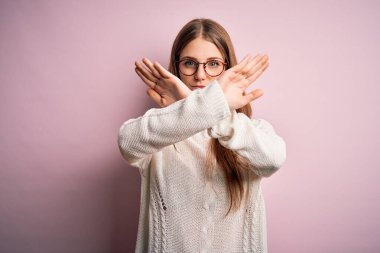 Genç, güzel, kızıl saçlı bir kadın pembe arka planda gözlük ve süveter takıyor.