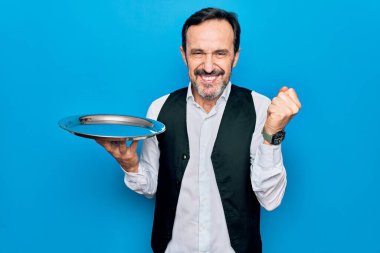 Orta yaşlı, yakışıklı garson, mavi arka planda duran tepside durmuş gururla bağırıyor, zaferi ve başarıyı kutluyor.