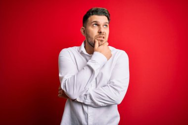 Mavi gözlü, zarif gömlekli, kırmızı izole edilmiş arka planda duran genç bir iş adamı bir soru hakkında endişeli ve gergin bir şekilde düşünüyor.