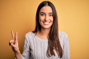 Sarı arka planda beyaz süveter giyen genç, güzel esmer kadın iki numarayı gösterirken kendinden emin ve mutlu bir şekilde gülümsüyor..