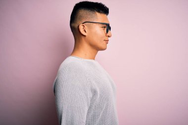 Pembe arka planda günlük süveter ve gözlük takan genç, yakışıklı Latin adam yan yana bakıyor, rahat poz veriyor, kendine güvenen bir gülümsemesi var..