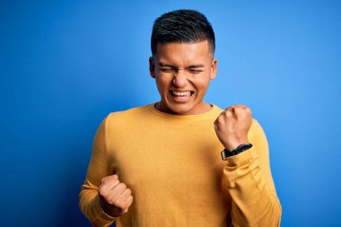 İzole edilmiş mavi arka plan üzerinde sarı günlük süveter giyen genç, yakışıklı latin adam kolları havada ve gözleri kapalı olarak başarının şaşkınlığını ve şaşkınlığını kutluyor. Kazanan konsept.