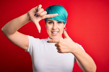 Mavi saçlı, kırmızı arka planda sıradan tişörtler giyen, gülümseyen, elleri ve parmakları mutlu yüzlü, çerçeveli genç, güzel bir kadın. Yaratıcılık ve fotoğrafçılık kavramı.