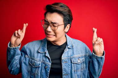 Kırmızı arka plan üzerinde kot ceket ve gözlük takan yakışıklı Çinli genç adam umut ve gözler kapalı gülümseyerek el işareti yapıyor. Şans ve batıl inanç..