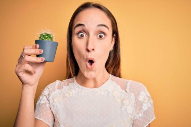 Güzel bir kadın, elinde küçük bir kaktüs saksısı ile izole edilmiş sarı arka plan üzerinde şaşkınlıktan korkmuş ve korku ifadesinden korkan genç bir kadın.