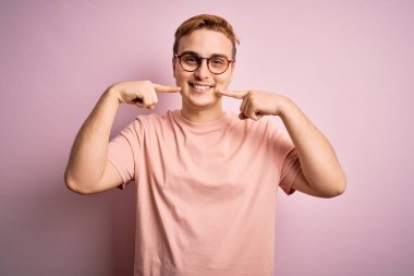 Genç, yakışıklı, günlük tişört giyen, pembe arka planda duran, neşeli gülümseyen, parmakları, dişleri ve ağzı olan bir adam. Diş sağlığı konsepti.