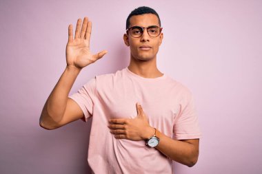 Yakışıklı Afro-Amerikalı adam pembe arka plan üzerine günlük tişört ve gözlük takıyor. Göğüs ve avuç içlerine kadar yemin ediyor. Sadakat yemini ediyor.