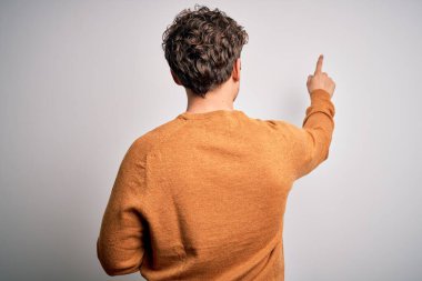 Sarışın, kıvırcık saçlı, beyaz arka planda sıradan bir süveter giyen, parmak uçlarıyla öne doğru poz veren genç, yakışıklı bir adam.