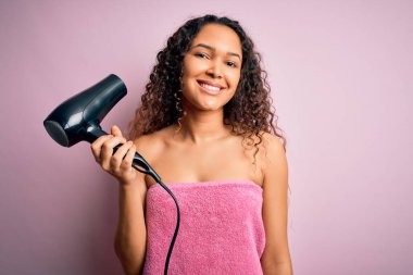 Kıvırcık saçlı güzel bir kadın banyo üstüne duş havlusu giyer, saç kurutma makinesi kullanır, güler yüzlü, kendine güvenen bir gülümsemeyle dişlerini gösterir.