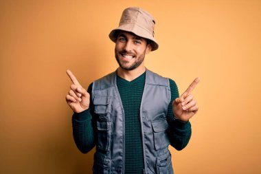 Tatilde sakallı yakışıklı turist sarı arka planda kaşif şapkası takıyor ve parmaklarıyla farklı yönleri işaret ederek gülümsüyor. Reklam için boşluk kopyala