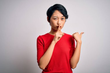 Genç, güzel Asyalı bir kız, günlük kırmızı tişörtler giyip izole edilmiş beyaz arka planda dikiliyor ve parmaklarını yan tarafı işaret ederek sessiz olmasını istiyor. Sessizlik ve gizli konsept.