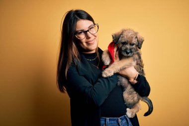 Güzel, esmer bir kadın, sevimli bir köpek yavrusunu izole edilmiş sarı arka planda tutuyor. Akıllı yüzünde kendine güvenen bir ifade var ve ciddi düşünüyor.