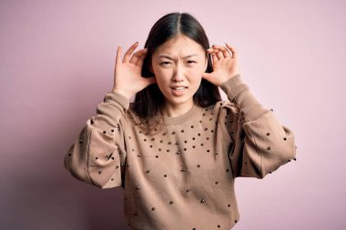Genç, güzel Asyalı bir kadın pembe arka planda şık bir süveter giyiyor. İki elini de kulak hareketi yaparken duymaya çalışıyor. Dedikoduya meraklı. İşitme problemi, sağır