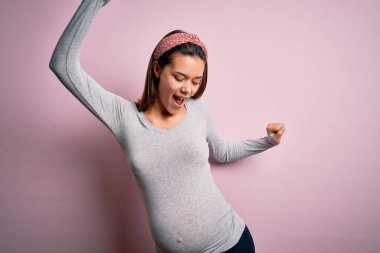 Genç, güzel, hamile bir kız pembe arka planda bebek bekliyor mutlu ve neşeli dans ediyor, gülümsüyor rahat ve kendinden emin bir şekilde müzik dinliyor.