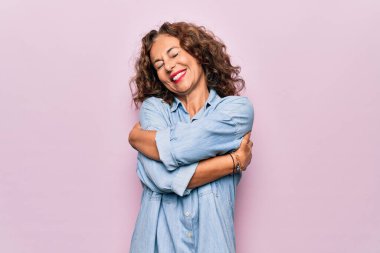 Orta yaşlı güzel bir kadın pembe arka planda sıradan bir kot gömlek giyip kendine sarılıyor mutlu ve pozitif, kendinden emin gülümsüyor. Sevgi ve özgüven.