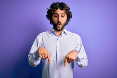 Mor arka plan üzerinde duran sakallı, genç ve yakışıklı iş adamı parmaklarını göstererek reklamını yapıyor, şaşırmış yüzünü ve ağzını açıyor.