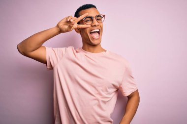 Yakışıklı Afro-Amerikan erkeği pembe arka planda günlük tişört ve gözlük takıyor. Yüzünde parmaklarıyla barış sembolü yapıyor. Gülümsüyor, zafere gülümsüyor.
