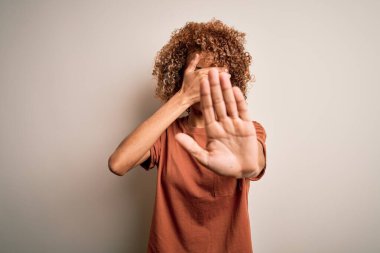 Kıvırcık saçlı güzel Afro-Amerikalı kadın beyaz arka plan üzerine günlük tişört giyip gözleri ellerle kapatıyor ve üzgün ve korku ifadesiyle dur hareketi yapıyor. Utanç verici ve olumsuz bir kavram..