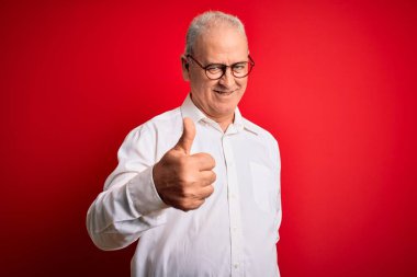 Orta yaşlı yakışıklı, beyaz gömlekli ve kırmızı arka planda gözlüklü bir adam mutlu bir el hareketi yapıyor. Kameraya bakıp başarıyı gösteren ifade onaylanıyor.