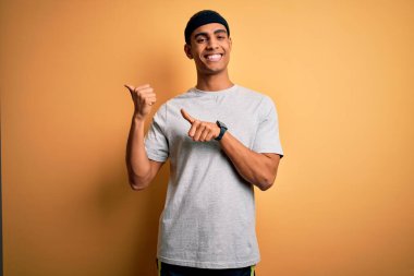 Yakışıklı Afro-Amerikan sporcu sarı arka planda spor giysisi giyiyor. Arka tarafı işaret ediyor. Elleri ve başparmakları havada, kendinden emin gülümsüyor.