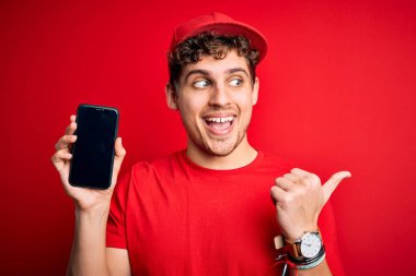 Kıvırcık saçlı, şapkalı, kırmızı arka planda akıllı telefon tutan ve gülümseyen yüzüyle baş parmağıyla yüzünü gösteren genç bir teslimatçı.