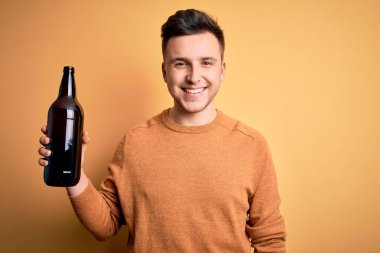 Genç, yakışıklı, beyaz bir adam sarı arka planda bir şişe bira içiyor. Mutlu bir yüzü var. Kendine güvenen bir gülümsemeyle dişlerini gösteriyor.