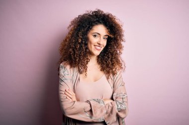 Kıvırcık saçlı, piercingli, sıradan pembe tişörtlü mutlu yüzlü, kollarını kavuşturup kameraya bakan güzel bir kadın. Pozitif kişi.