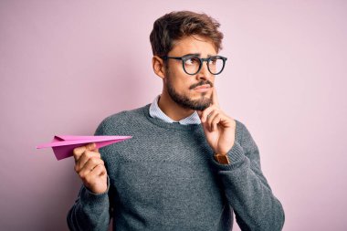 Elinde kağıt uçak tutan, pembe arka planda gözlük takan yakışıklı genç bir adam. Ciddi bir yüz. Soru soruyor, kafası karışık bir fikir.
