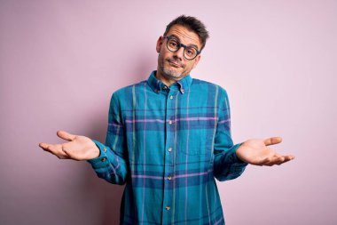 Sıradan gömlek ve gözlük takan genç yakışıklı bir adam soyutlanmış pembe arka plan üzerinde duruyor. Kolları ve elleri havada, kafası karışmış bir ifade. Şüphe kavramı.