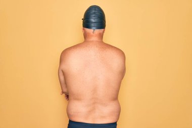 Orta yaşlı, yaşlı, gri saçlı, mayo, kep ve gözlük takan, kollarını kavuşturup arkaya bakan bir yüzücü.